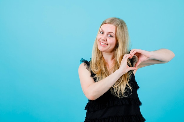 La ragazza bionda sorridente sta facendo il gesto del cuore su sfondo blu