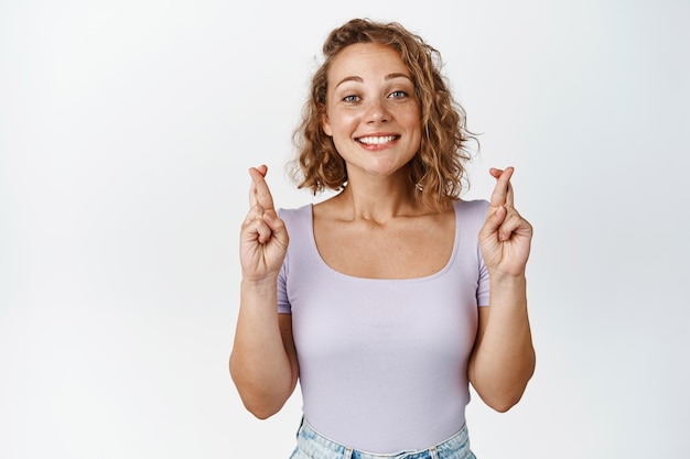 La ragazza bionda speranzosa incrocia le dita per buona fortuna, in attesa di qualcosa con eccitazione, sfondo bianco.