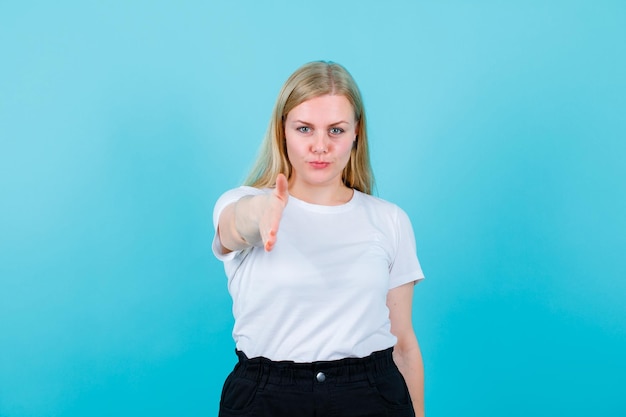 La ragazza bionda sta mostrando ciao gesto estendendo la mano alla fotocamera su sfondo blu