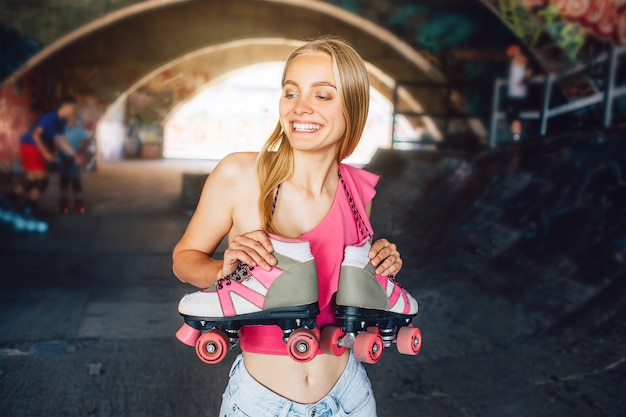 La ragazza bionda sta sorridendo. È in piedi in una sala di addestramento per pattini a rotelle. I rulli sono appesi al collo. Li tiene con le mani. La ragazza sorride e guarda a sinistra.