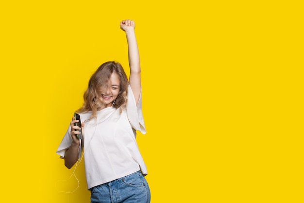 La ragazza bionda sta tifando per un muro giallo con spazio libero mentre ascolta la musica sugli auricolari e alza la mano