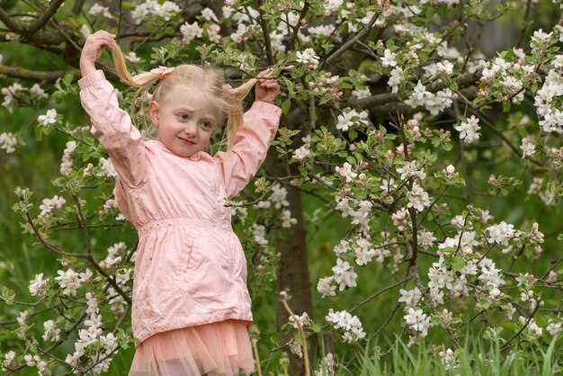 La ragazza bionda tra gli alberi in fiore si siede sull'erba la ragazza con due code accanto all'albero in fiore primaverile in natura primaverile