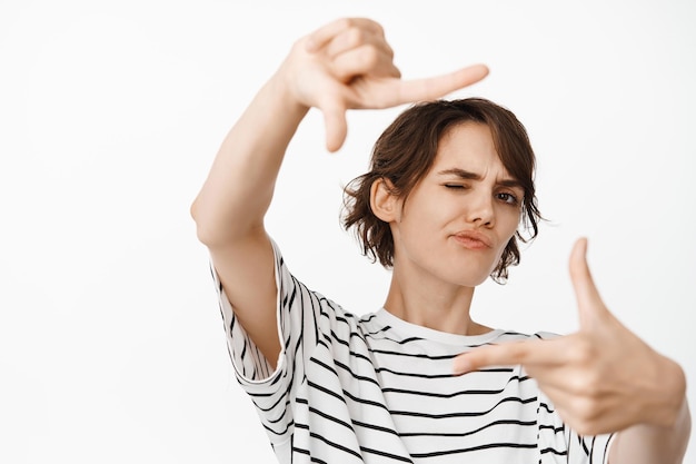 La ragazza bruna creativa guarda attraverso la cornice della mano premurosa, pensa alla scena delle riprese, scatta foto, in piedi su sfondo bianco