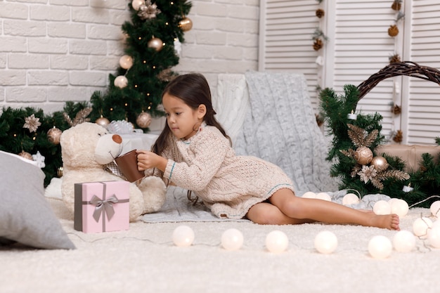 La ragazza carina beve cioccolata calda e gioca con un orsacchiotto a casa per Natale