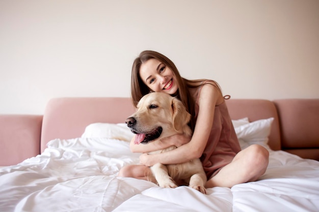 La ragazza carina in pigiama si siede sul letto e abbraccia la donna del cane di razza golden retriever al mattino