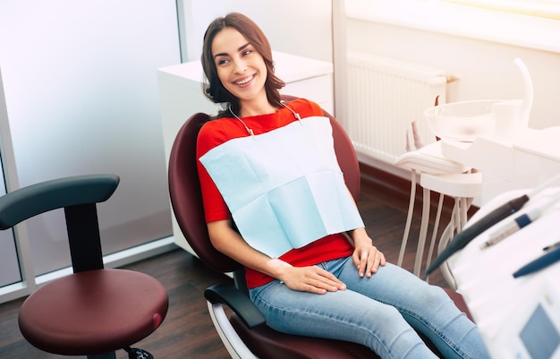 La ragazza carina magra vestita di maglione rosso e jeans denim è seduta sulla poltrona del dentista e aspetta il dottore con tutte le piacevoli emozioni