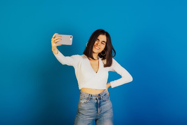 La ragazza carina sorridente divertente strizza l'occhio mentre prende selfie sul suo telefono isolato su una superficie blu.