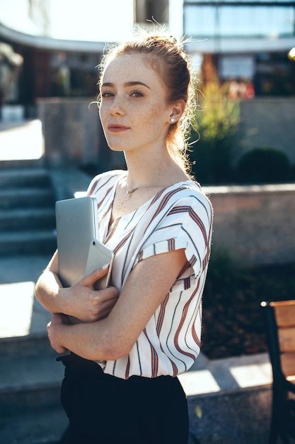 La ragazza caucasica dai capelli rossi con le lentiggini sta esaminando la macchina fotografica mentre tiene un computer portatile e un cellulare