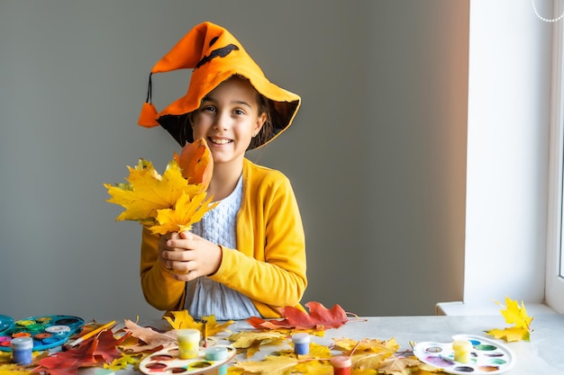 La ragazza caucasica in costume da strega si siede al tavolo di casa e prepara l'artigianato per le vacanze di Halloween. Fai da te e arredamento in camera di carta.