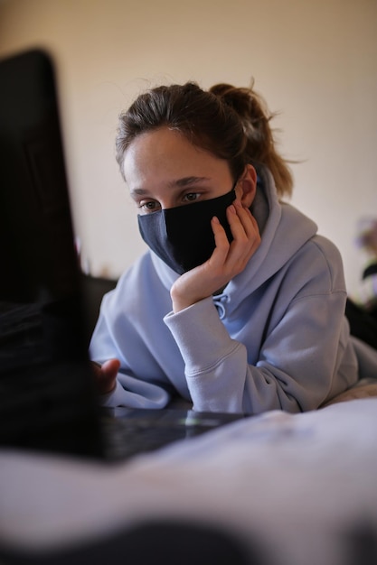 La ragazza caucasica in felpa con cappuccio blu e maschera nera lavora su un laptop a casa isolata