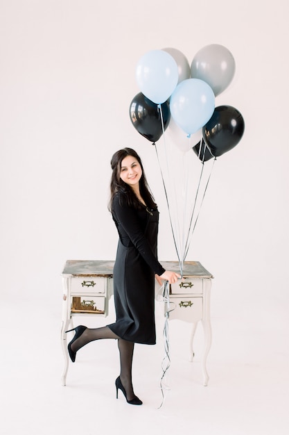 La ragazza caucasica positiva felice in vestito nero tiene il mazzo di aerostati, stante vicino alla tavola bianca d'annata con i cassetti, aspetta gli ospiti, isolato sopra fondo bianco. Concetto di festa di compleanno