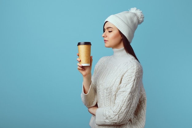 La ragazza che indossa un cappello bianco e un maglione si diverte a bere il caffè dalla tazza da asporto