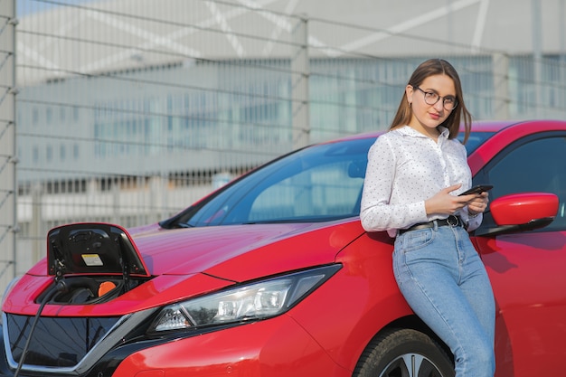 La ragazza che per mezzo dello Smart Phone e l'attesa dell'alimentazione elettrica si collegano ai veicoli elettrici per caricare la batteria in automobile. Auto ecologica collegata e ricarica batterie