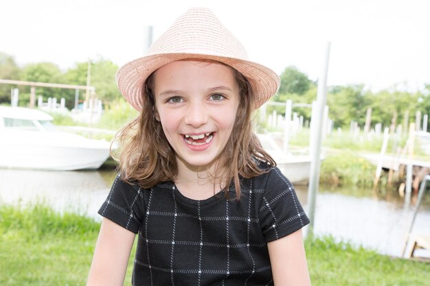 La ragazza che ride con il cappello di paglia rosa sorride e la giornata felice nel parco lungo il fiume