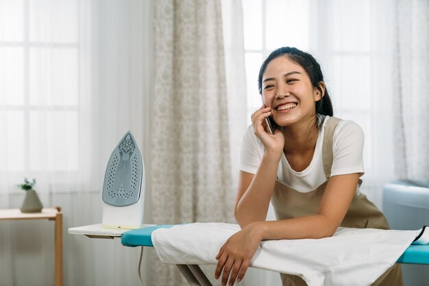 la ragazza cinese felice che si appoggia sull'asse da stiro sorride mentre si chatta sul telefono cellulare in un interno luminoso della lavanderia domestica.