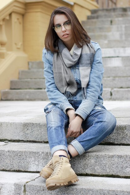 La ragazza con capelli scuri sta sedendosi sulle scale e sta esaminando la macchina fotografica. Occhiali e jeans