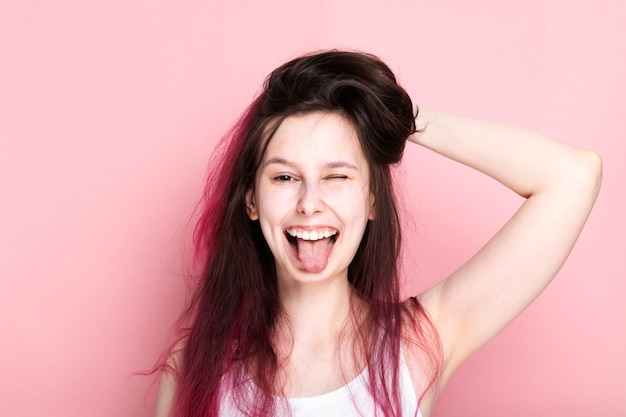 La ragazza con i capelli arruffati rosa senza trucco fa la faccia divertente e attacca la sua lingua su fondo rosa