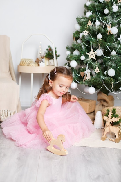 La ragazza con i capelli biondi con un vestito rosa elegante vicino all'albero di Natale si siede sul pavimento
