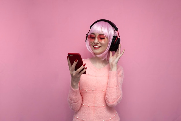 La ragazza con i capelli rosa in cuffia ascolta musica e tiene uno smartphone hipster utilizza un telefono su uno sfondo rosa