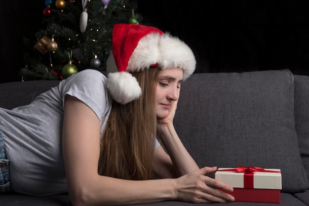 La ragazza con il cappello di Babbo Natale guarda pensierosa il regalo sdraiato sul divano sullo sfondo di un albero di natale. Anticipazione della vacanza.