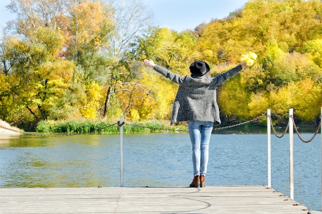 La ragazza con il cappello è in piedi sul molo con le mani alzate.