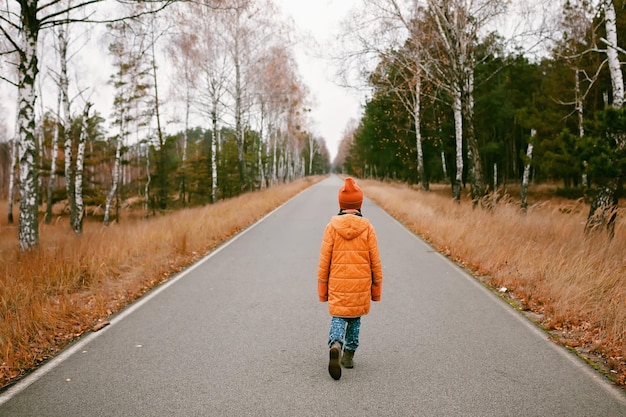 La ragazza con il cappotto arancione cammina lungo la strada nel pensiero della foresta autunnale e nelle vibrazioni autunnali della salute mentale