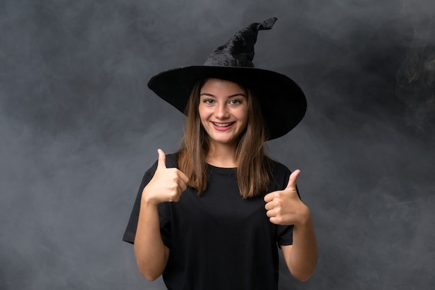 La ragazza con il costume della strega per le feste di Halloween sopra la parete scura isolata dando pollici aumenta il gesto