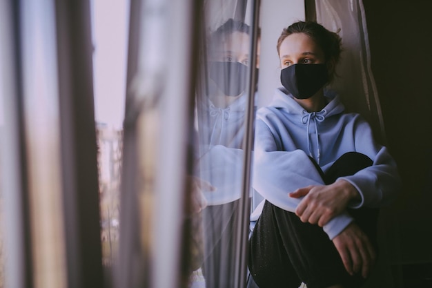 La ragazza con la maschera nera è in piedi alla finestra di casa in isolamento in elaborazione di foto vintage