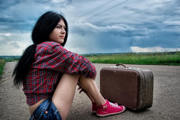 La ragazza con la valigia vintage si siede sull'auto in attesa dell'asfalto. Ragazza che viaggia in autostop.