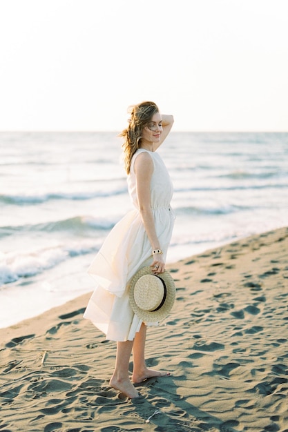 La ragazza con un cappello di paglia si trova sulla spiaggia con i capelli che volano