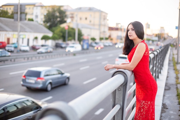 La ragazza con un lungo vestito rosso è in piedi vicino al recinto della carreggiata.