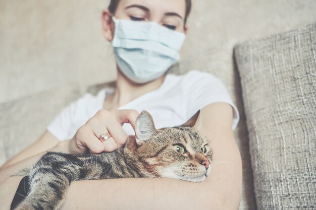 La ragazza con una mascherina medica è auto-isolata a casa e sta riposando con un gatto sul divano.