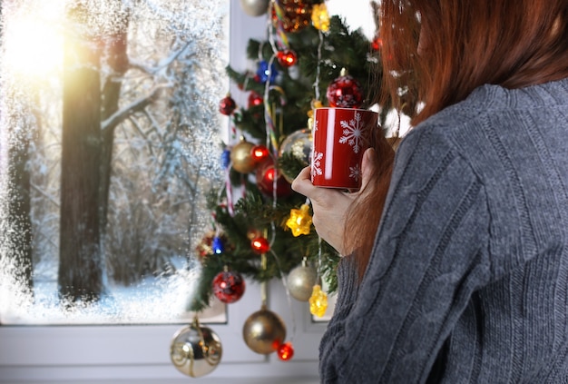 La ragazza con una tazza di bevanda calda sta davanti a una finestra a Capodanno