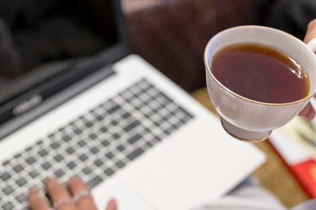 La ragazza con una tazza di tè in mano sta scrivendo su un computer portatile