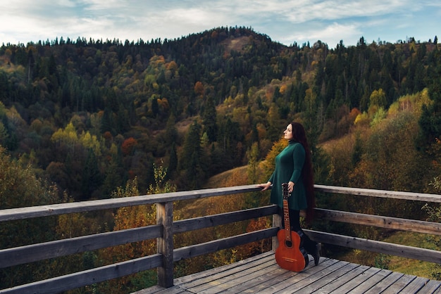La ragazza dai capelli lunghi tiene la chitarra, in piedi vicino al parapetto in montagna in autunno.