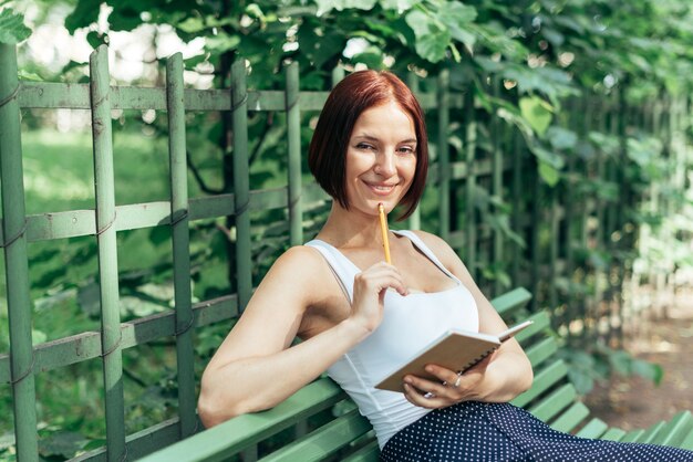 La ragazza dai capelli rossi caucasica si siede in estate nel parco su una panchina e si rilassa tenendo un blocco note.
