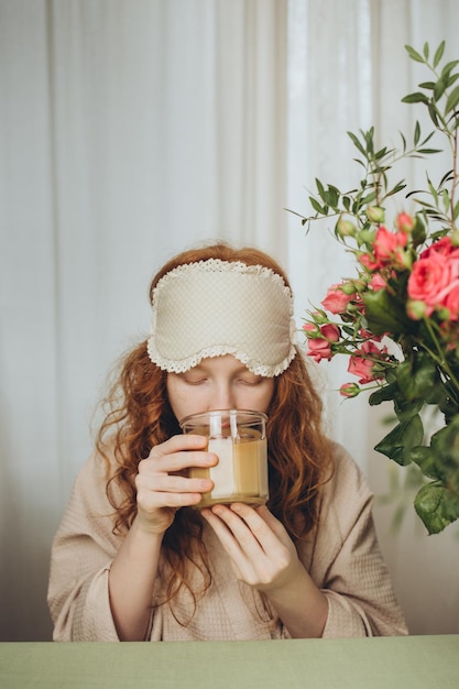 La ragazza dai capelli rossi con gli occhi chiusi beve il caffè del mattino da una tazza di vetro al tavolo