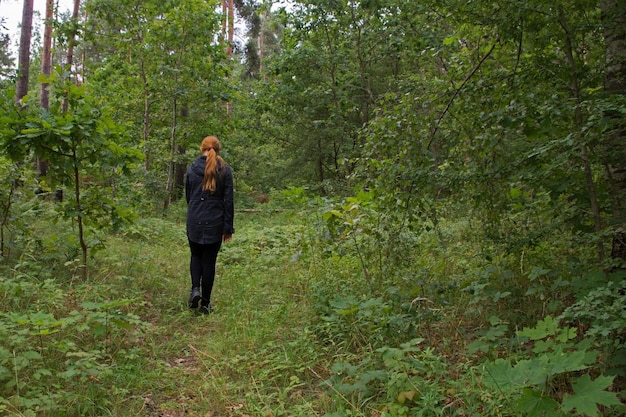 La ragazza dai capelli rossi in giacca nera e leggings sta camminando lungo il sentiero sull'erba verde nella foresta