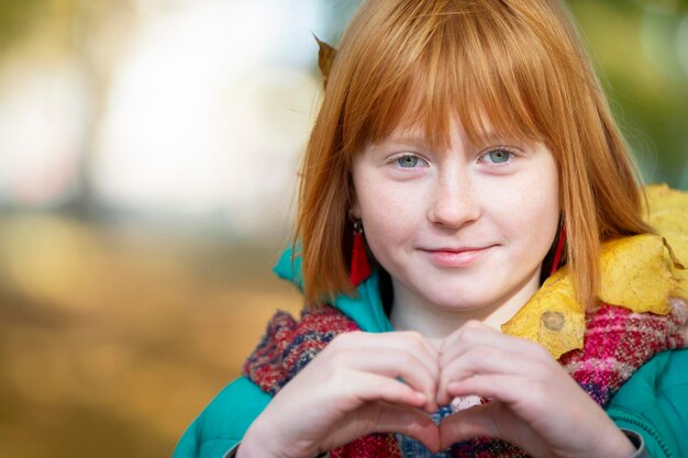 La ragazza dai capelli rossi nel parco autunnale mostra un segno di cuore