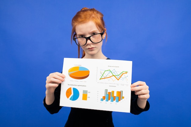 La ragazza dai capelli rossi seria dell'adolescente in vetri in un vestito nero mostra gli strati con i grafici e le statistiche blu