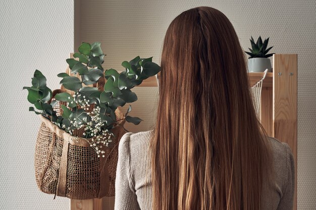 La ragazza decora uno scaffale in legno con una borsa ecologica con rami di eucalipto, fiori, piante d'appartamento