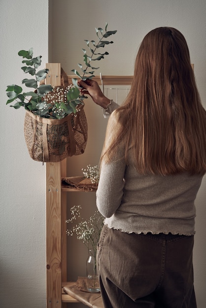 La ragazza decora uno scaffale in legno con una borsa ecologica con rami di eucalipto, fiori, piante d'appartamento