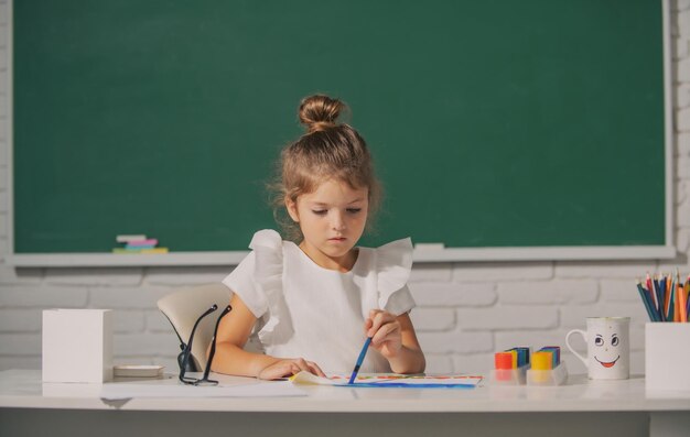 La ragazza del bambino disegna in classe seduta a un tavolo divertendosi sulla lavagna della scuola sfondo divertente ki