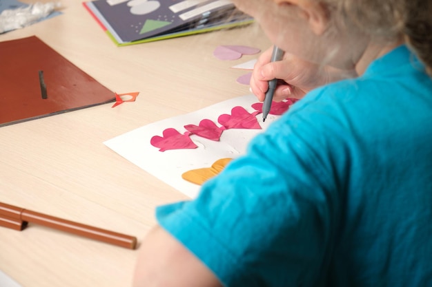 La ragazza del bambino in età prescolare in maglietta blu fa applique seduta al tavolo, cuori ritagliati di bougue colorata per la carta per San Valentino, creatività dei bambini, dermatite atopica sulle mani di un bambino