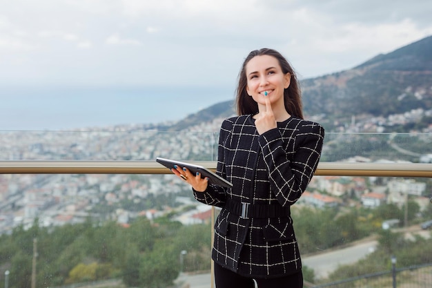 La ragazza del blogger del computer di Planshet sta pensando tenendo l'indice sul mento sullo sfondo della vista sulla città