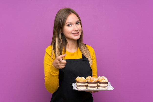 La ragazza dell'adolescente che tiene i lotti di mini mini torte differenti sopra la parete viola isolata indica il dito voi