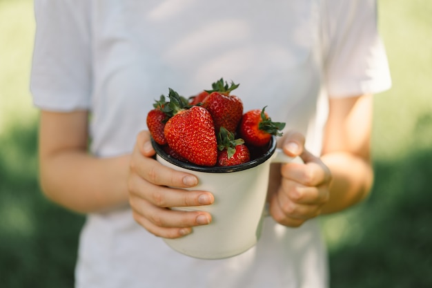 La ragazza dell'adolescente tiene in mano una gustosa fragola brillante matura in una tazza di cibo vitaminico estivo ma...