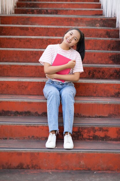 La ragazza della scuola indossa una camicia rosa e un jeans che tiene e tiene in mano un libro rosa sulle scale