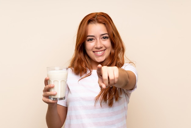 La ragazza della testarossa dell'adolescente che tiene un bicchiere di latte sopra fondo isolato indica il dito voi con un'espressione sicura