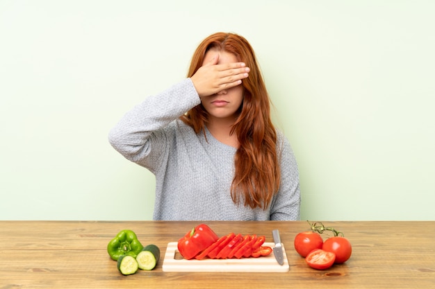 La ragazza della testarossa dell'adolescente con le verdure in una copertura della tabella osserva a mano